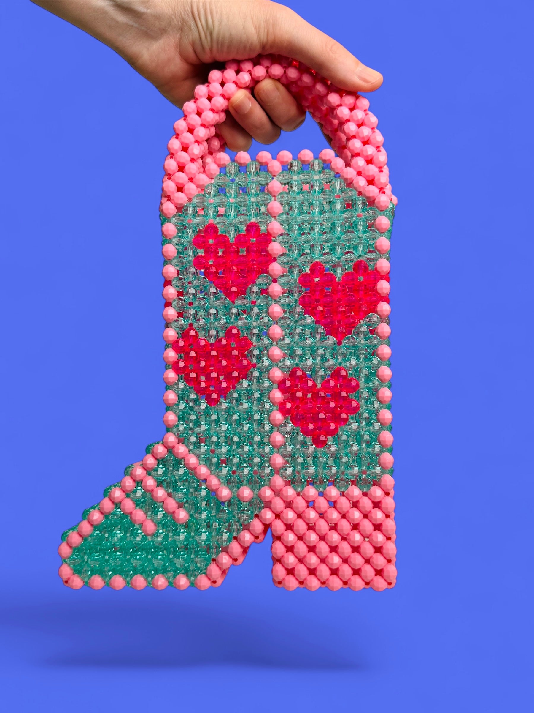 A hand holds a beaded handbag shaped like a cowboy boot, featuring an aqua green body with pink and red beaded hearts, pink beaded trim, and a pink beaded handle, set against a solid purple background.