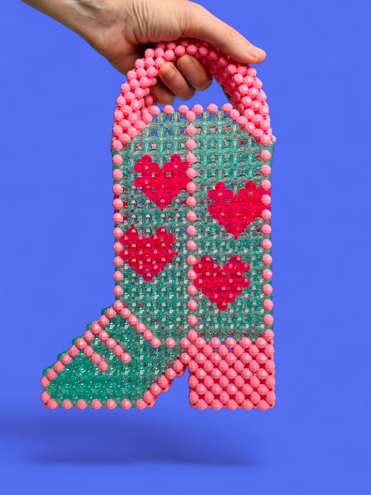 A hand holds a beaded handbag shaped like a cowboy boot, featuring an aqua green body with pink and red beaded hearts, pink beaded trim, and a pink beaded handle, set against a solid purple background.
