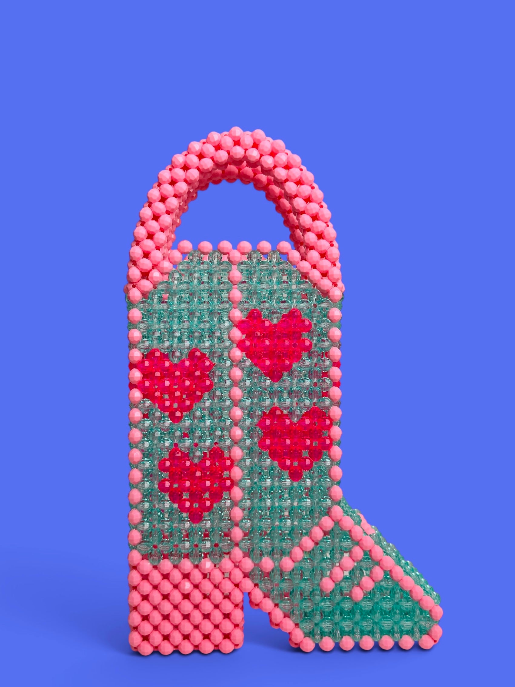 A hand holds a beaded handbag shaped like a cowboy boot, featuring an aqua green body with pink and red beaded hearts, pink beaded trim, and a pink beaded handle, set against a solid purple background.