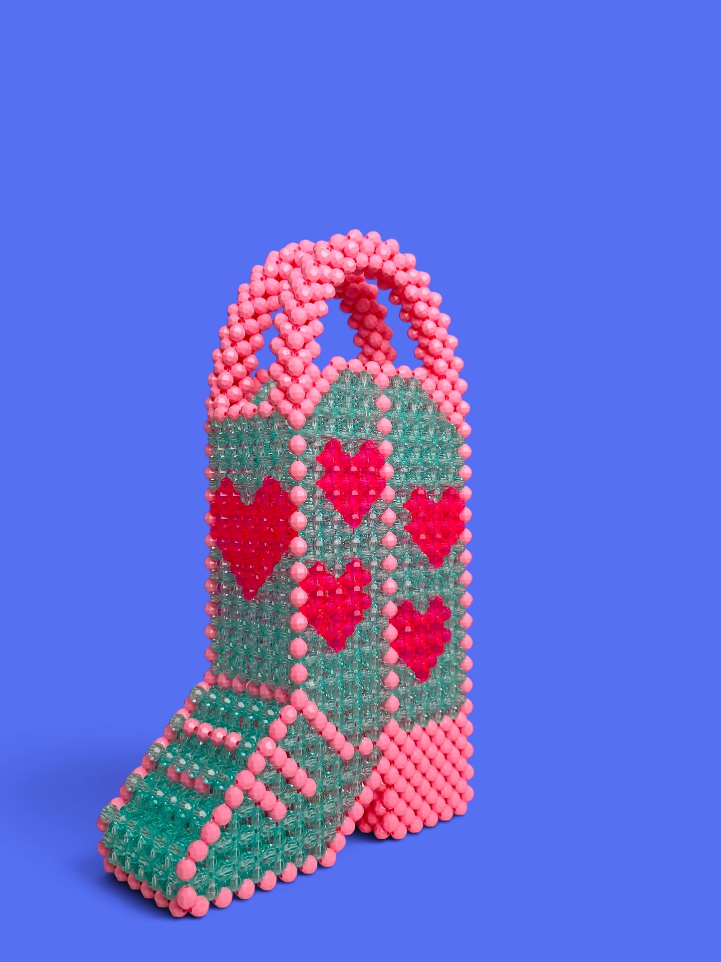 A hand holds a beaded handbag shaped like a cowboy boot, featuring an aqua green body with pink and red beaded hearts, pink beaded trim, and a pink beaded handle, set against a solid purple background.